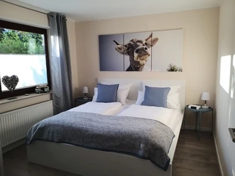 Schlafzimmer 1 Gemütliches Schlafzimmer mit Doppelbett, blau-weißer Bettwäsche, Kuhbild über dem Bett, Tageslicht.