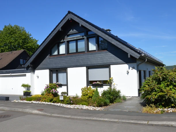 Außenansicht Weißes Einfamilienhaus mit Dachschindeln und Garten, gelegen in ruhiger Wohngegend bei Sonnenschein.