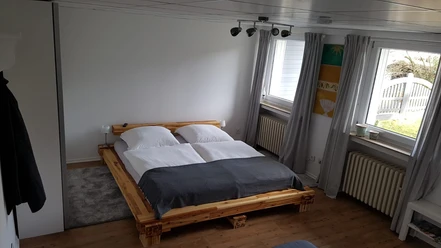 Ferienwohnung Oskar Helles Schlafzimmer mit Doppelbett, grauen Vorhängen und Holzboden, Blick auf den Garten.
