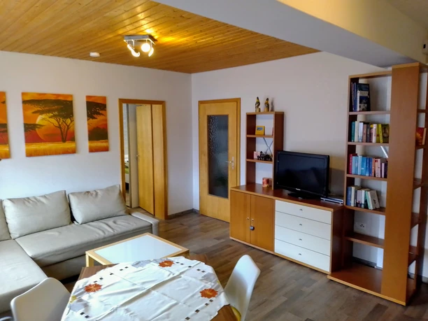 Wohnzimmer Gemütliches Wohnzimmer mit beiger Sofagarnitur, Holzmöbeln, Bücherregal und warm beleuchteter Holzdecke.