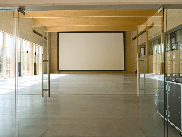 Halle Großer, leerer Raum mit Glaswänden und Holzelementen, im Hintergrund eine Projektionsfläche.