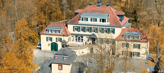 Schloss Eulenbroich Luftaufnahme eines historischen gelben Herrenhauses mit roten Dächern, umgeben von herbstlichen Bäumen.