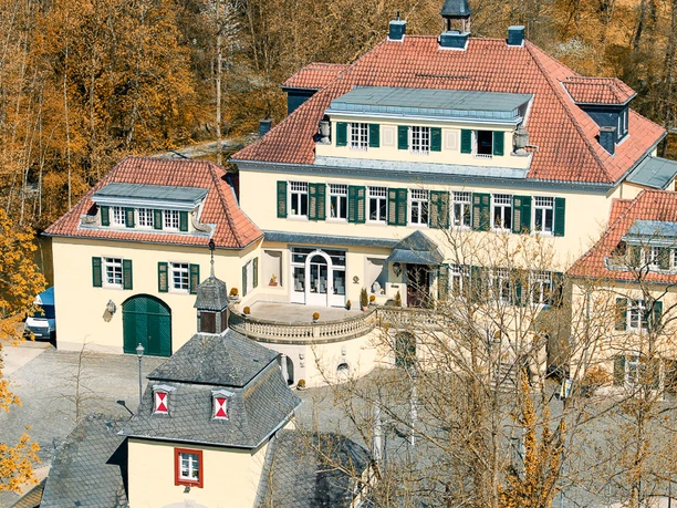 Schloss Eulenbroich Luftaufnahme eines historischen gelben Herrenhauses mit roten Dächern, umgeben von herbstlichen Bäumen.