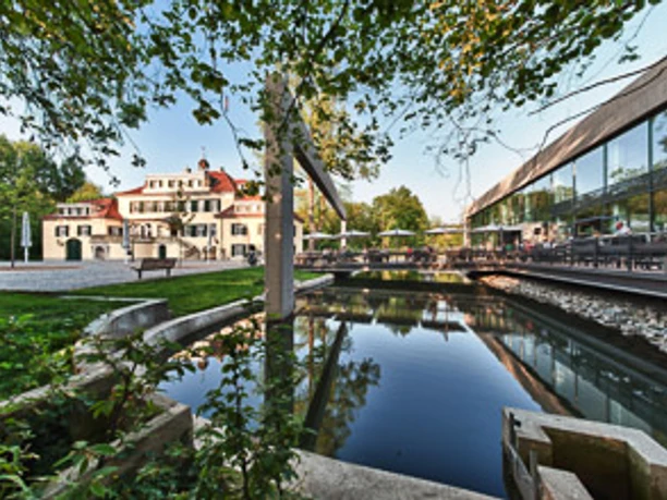 Schloss Eulenbroich Ein elegantes Herrenhaus mit rotem Dach neben einem modernen Glasgebäude spiegelt sich in einem Teich.