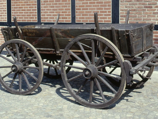 Fuhrmannswagen Ein alter, hölzerner Pferdewagen mit vier großen Rädern steht auf einem Kopfsteinpflaster vor einer Backsteinmauer.