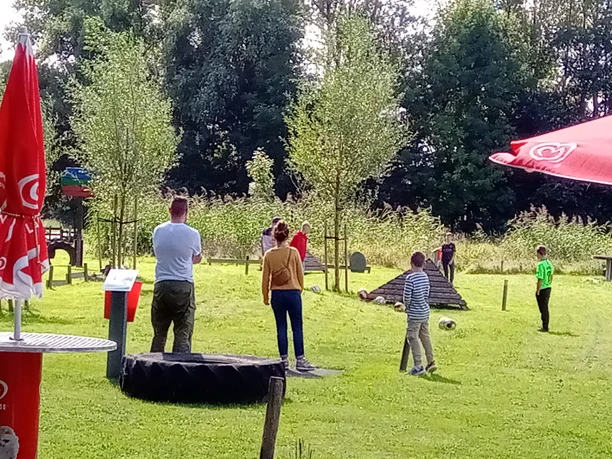 Activity Park FussBallGolf - Bahnen Besucher spielen Fußballgolf auf einer grünen Anlage mit Holzhindernissen und roten Sonnenschirmen.
