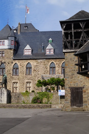 Schloss Burg Burganlage aus Stein mit Zinnenturm, Statue im Vordergrund, blauer Himmel im Hintergrund.