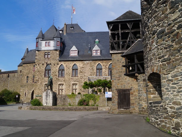 Schloss Burg Burganlage aus Stein mit Zinnenturm, Statue im Vordergrund, blauer Himmel im Hintergrund.