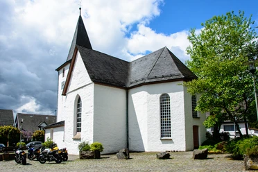 Bonte Kerke Lieberhausen Weiß verputzte Kirche mit spitzem Turm, umgeben von Bäumen und einigen parkenden Motorrädern.