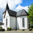 Bonte Kerke Lieberhausen Weiß verputzte Kirche mit spitzem Turm, umgeben von Bäumen und einigen parkenden Motorrädern.