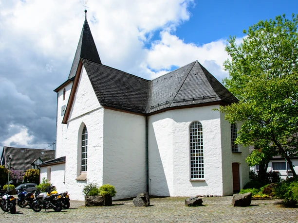 Bonte Kerke Lieberhausen Weiß verputzte Kirche mit spitzem Turm, umgeben von Bäumen und einigen parkenden Motorrädern.