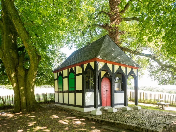 Rochuskapelle in Sand Ein malerisches Fachwerkhäuschen unter einem majestätischen Baum auf einer lichtdurchfluteten Wiese.