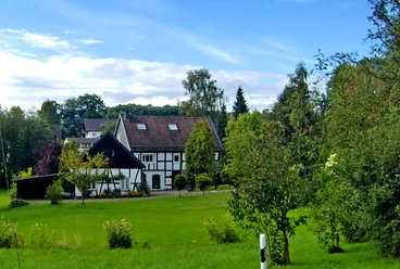 Fachwerk in Reichshof-Sinspert Fachwerkhaus mit rotem Ziegeldach in grüner Landschaft, umgeben von Bäumen und Wiese unter blauem Himmel.