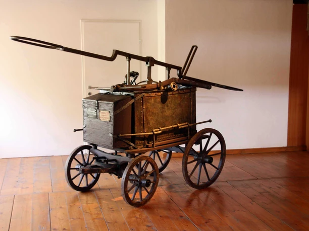 Bild Historische Handfeuerspritze aus Holz mit großen Rädern und langen Griffen auf Holzboden.
