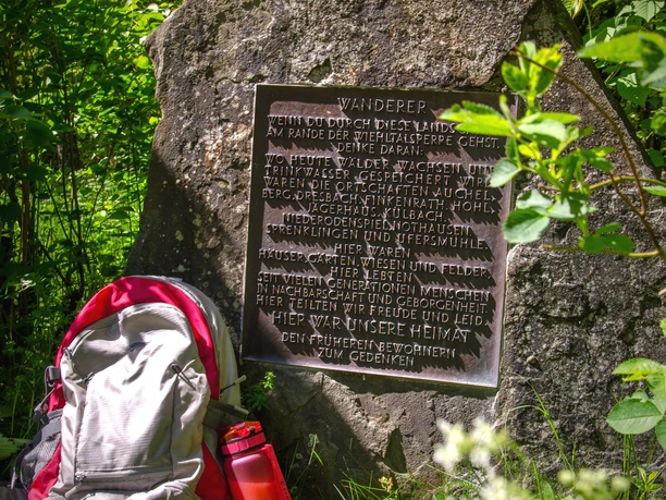 Wiehlquellen-Wanderweg Gedenkstein im Wald mit Inschrift, umgeben von grünen Büschen; Rot-weißer Rucksack und Schuhen davor.