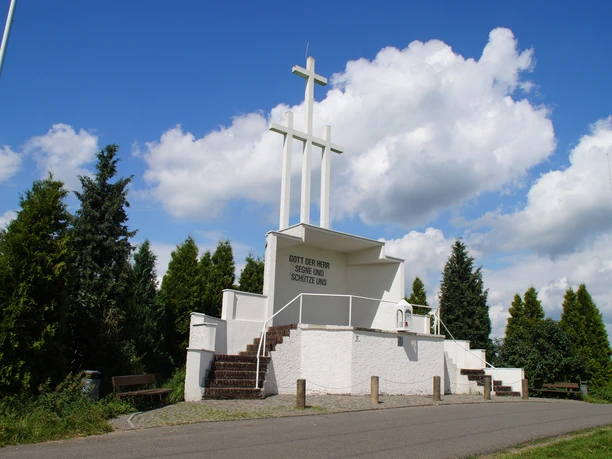 Freiluftaltar Ommerborn Drei weiße Kreuze auf einem Podest in einer grünen Umgebung unter blauem Himmel mit Wolken.