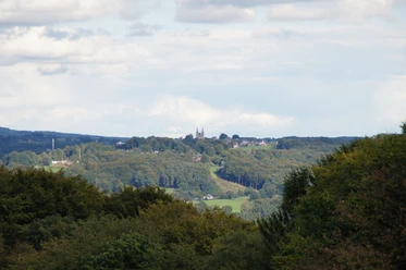 Blick auf Marialinden Landschaft mit bewaldeten Hügeln, im Hintergrund eine Kirche auf einem Hügel unter leicht bewölktem Himmel.