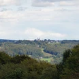 Blick auf Marialinden Landschaft mit bewaldeten Hügeln, im Hintergrund eine Kirche auf einem Hügel unter leicht bewölktem Himmel.