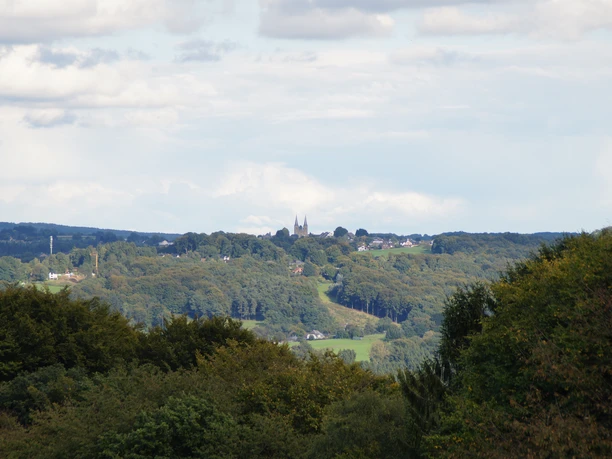 Blick auf Marialinden Landschaft mit bewaldeten Hügeln, im Hintergrund eine Kirche auf einem Hügel unter leicht bewölktem Himmel.