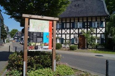 Infotafel am Obstweg Ein traditionelles Fachwerkhaus hinter einem Infoboard an einer sonnigen Straße in Leichlingen.