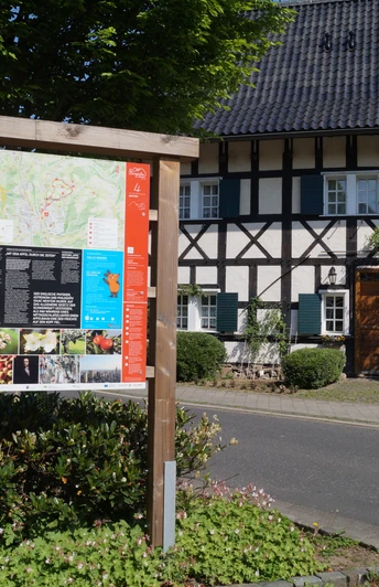 Infotafel am Obstweg Ein traditionelles Fachwerkhaus hinter einem Infoboard an einer sonnigen Straße in Leichlingen.