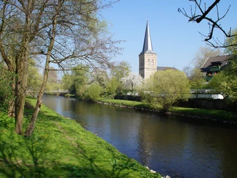 Impressionen vom Obstweg Kirche am Fluss in Leichlingen, umgeben von Bäumen und blühendem Grün unter klarem Himmel.