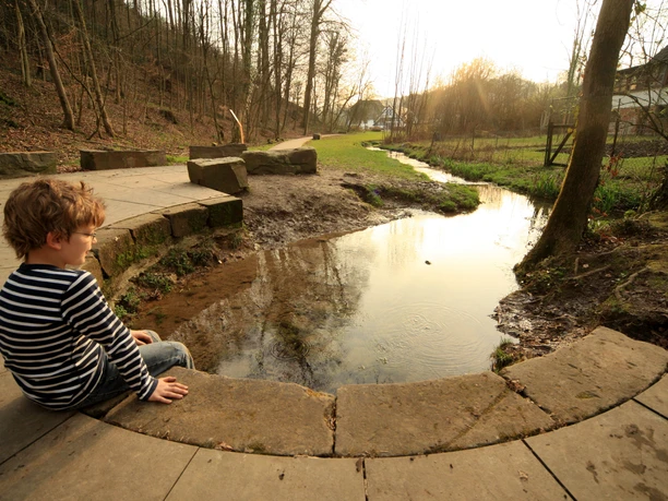Quelle der Strunde Ein Junge in gestreiftem Pullover sitzt auf einer Steinmauer am Rand eines kleinen Baches im Wald.