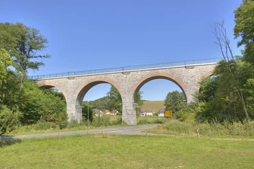 Lindlar - Linde - Bruch Viadukt Steinviadukt mit fünf Bögen, umgeben von Bäumen, mit Dorf und Hügel im Hintergrund unter blauem Himmel.