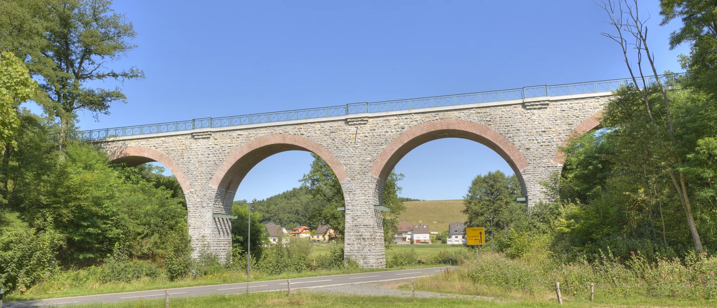 Lindlar - Linde - Bruch Viadukt Steinviadukt mit fünf Bögen, umgeben von Bäumen, mit Dorf und Hügel im Hintergrund unter blauem Himmel.