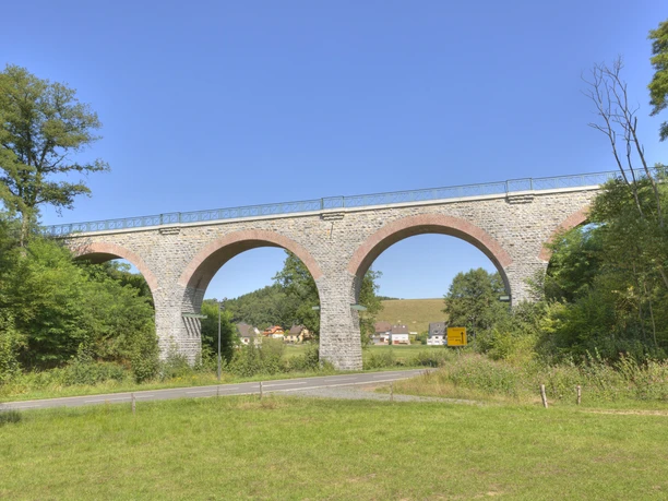 Lindlar - Linde - Bruch Viadukt Steinviadukt mit fünf Bögen, umgeben von Bäumen, mit Dorf und Hügel im Hintergrund unter blauem Himmel.