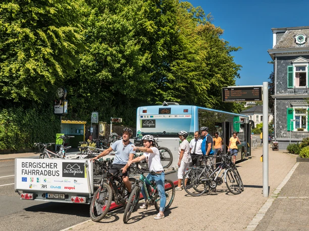 Bergischer FahrradBus, Haltestelle Hückeswagen Fahrradanhänger mit Rädern vor einem Bus; wartende Radfahrer beim Einsteigen in sonniger Stadtumgebung.