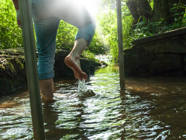 Naturtretbecken in Windfus Person watet barfuß durch einen klaren Bach, Sonnenlicht fällt durch üppige grüne Vegetation.