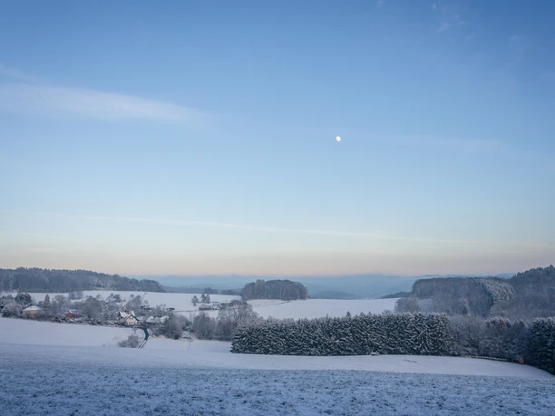 Lobscheid im Schnee Verschneite Landschaft in Oberösterreich, sanfte Hügel, Bäume, ein Dorf, blauer Himmel und Mond.