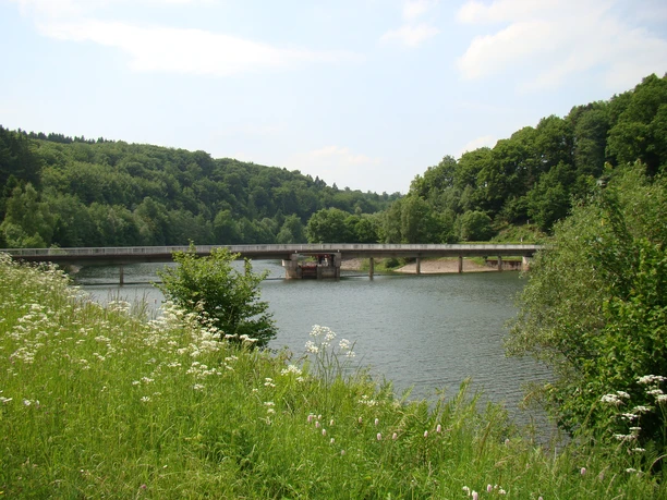 Impressionen vom Wasserweg Flusslandschaft mit Brücke, umgeben von grünen Wäldern und Wiesenblumen unter blauem Himmel.