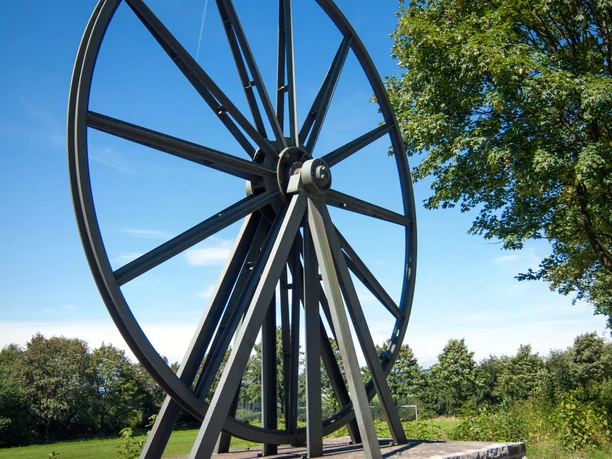 Wildberg Ein großes Rad aus Metall, auf einem steinernen Sockel montiert, steht im Grünen unter blauem Himmel.