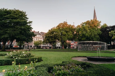 Parkanlage mit Blumenbeeten, Bäumen und Häusern im Hintergrund.
