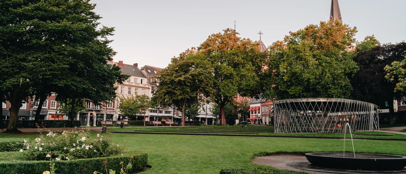 Parkanlage mit Blumenbeeten, Bäumen und Häusern im Hintergrund.