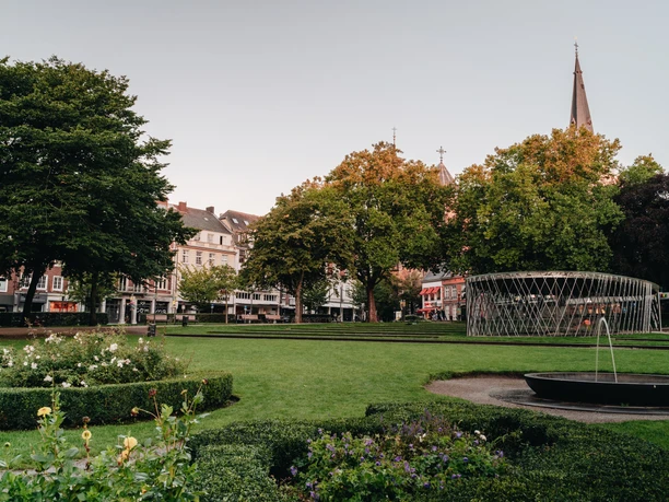 Parkanlage mit Blumenbeeten, Bäumen und Häusern im Hintergrund.