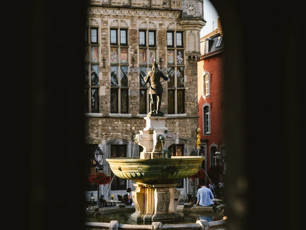 Karlsbrunnen Aachen von Rathaustreppen Blick durch einen Spalt auf die Bronzestatue von Karl dem Großen auf einem Brunnen mit der historischen Fassade des Aachener Rathauses im Hintergrund.