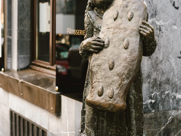Bronze-Figur eines Mädchens vor einem Schaufenster, das eine fast körpergroße, mit Mandeln verzierte Printe, ebenfalls aus Bronze, in den Händen hält.
