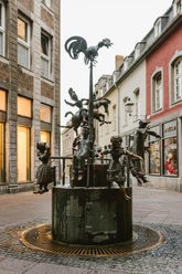 Brunnen mit verschiedenen Figuren aus Bronze, darunter Personen in altertümlicher Kleidung und ein Hahn ganz oben.