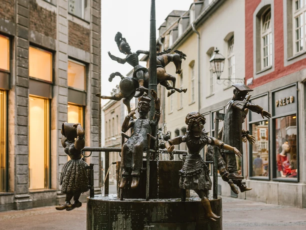 Brunnen mit verschiedenen Figuren aus Bronze, darunter Personen in altertümlicher Kleidung und ein Hahn ganz oben.