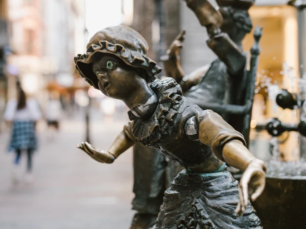 Bronzefigur einer Frau mit Haube und großen, runden Augen. Sie ist am Rand des Brunnens befestigt.