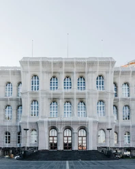 Historisches Bauwerk mit symmetrischen Fensterreihen, drei Eingangstüren und einer Fassade in hellem Grau. Es ist zurzeit mit einer netzartigen Schutzplane überzogen. Vor dem Gebäude erstreckt sich ein Platz.