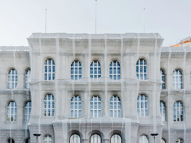 Historisches Bauwerk mit symmetrischen Fensterreihen, drei Eingangstüren und einer Fassade in hellem Grau. Es ist zurzeit mit einer netzartigen Schutzplane überzogen. Vor dem Gebäude erstreckt sich ein Platz.