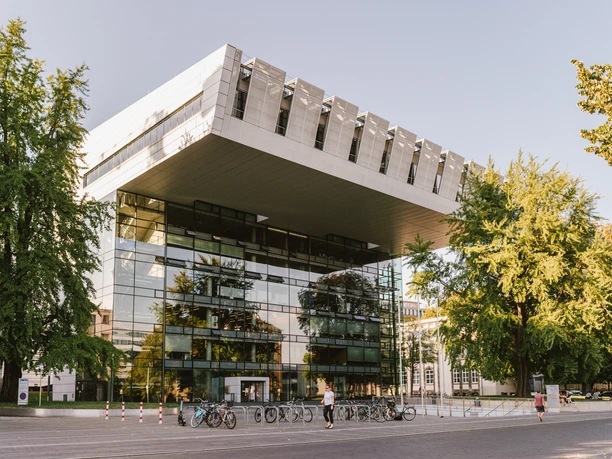 Modernes Gebäude in C-Form mit Glasfront und einem Vorplatz, umgeben von grünen Bäumen.