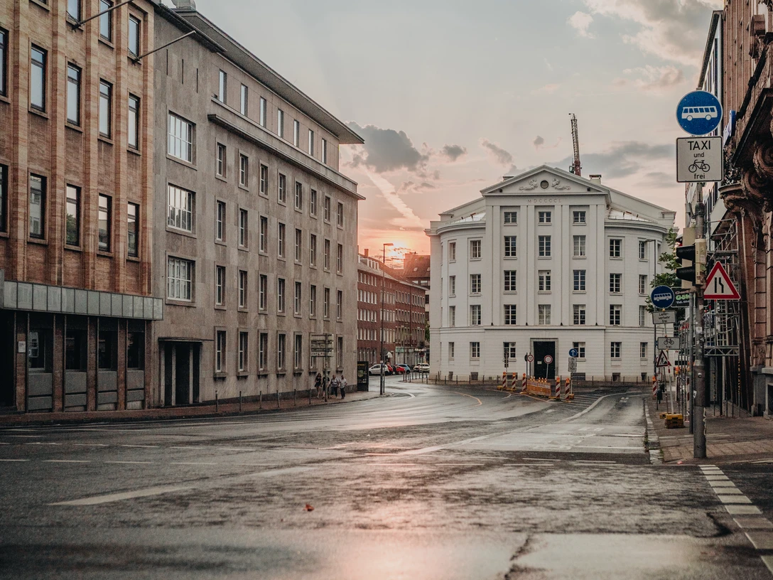 Theater Aachen Regen Blick über eine breite, nasse und von mehrstöckigen Gebäuden begleitete Straße auf das Theater Aachen und den Sonnenuntergang im Hintergrund.