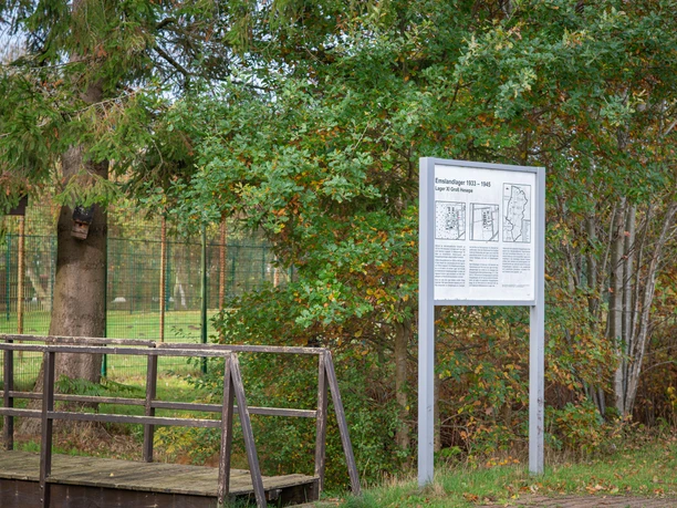 Emslandlager Groß Hesepe Infotafel zum Emslandlager Groß Hesepe vor Bäumen und Zaun im Grünen mit Holzsteg im Vordergrund.