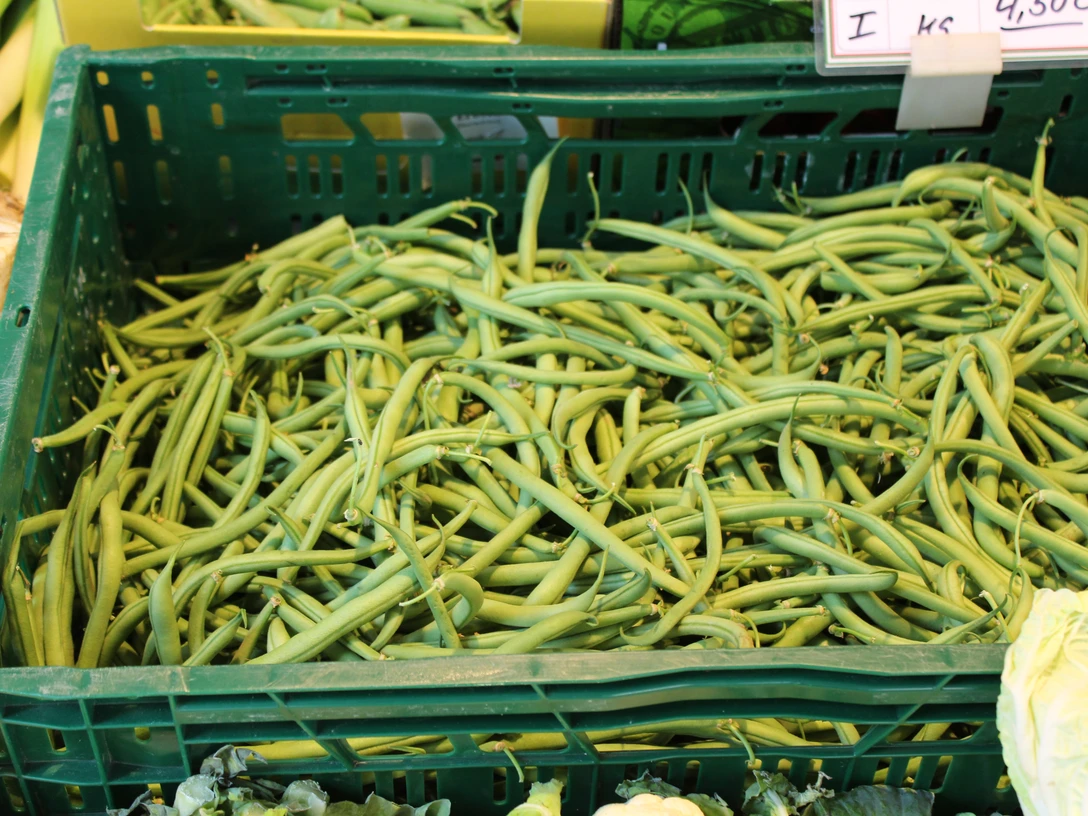 Frische grüne Bohnen in einer grünen Kunststoffkiste auf einem Wochenmarkt, umgeben von Gemüse.