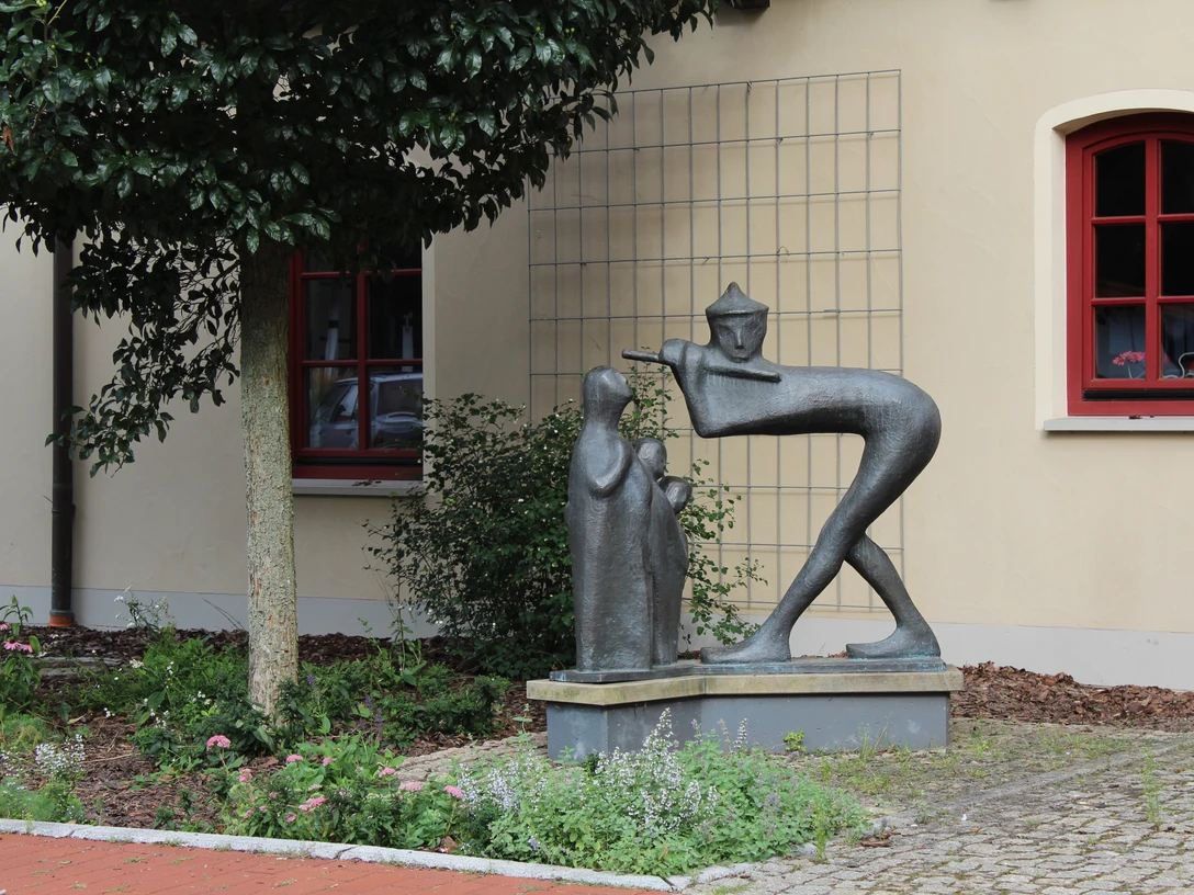 Bronzestatue eines Spielmanns mit Flöte und zuhörendem Kind vor einer beige Gebäude.